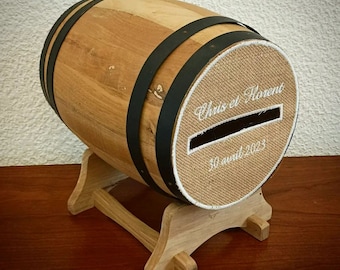 May include: A wooden barrel with black metal hoops and a burlap lid with a slot for money. The lid is embroidered with the text "Chris et Florent 30 avril 2023". The barrel is on a wooden stand.