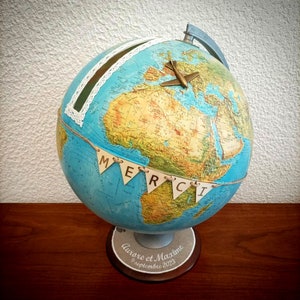Peut inclure: Un globe terrestre bleu avec une carte du monde, un petit avion au sommet et une bannière qui dit "MERCI". Le globe est sur un socle en bois avec le texte "Aurore et Maxime 9 septembre 2023".