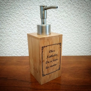 Könnte beinhalten: Ein Bambus-Seifenspender mit einer Edelstahlpumpe. Der Spender hat eine rechteckige Form und trägt den Text "Chez Nathalie, On se lave les mains!" in einem dekorativen Rahmen. Der Spender steht auf einer Holzoberfläche.