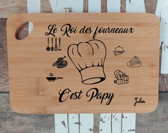 May include: A brown wooden cutting board with a handle. The board has a black design that says "Le Roi des fourneaux C'est Papy Jules". The design includes a chef's hat, a frying pan, a piece of meat, a cake, and a carrot.