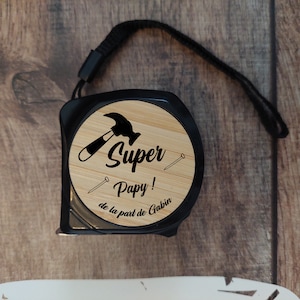 Può includere: Un metro a nastro nero e marrone con un viso in legno. Il viso ha un design a martello e chiodo nero e il testo "Super Pappy! de la part de Gabin".