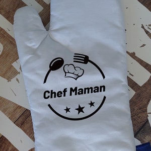 Könnte beinhalten: Ein weißer Topflappen mit blauem Saum. Er hat ein schwarzes Design mit einer Kochmütze, einem Löffel und einer Gabel mit dem Text "Chef Maman" und drei Sternen.