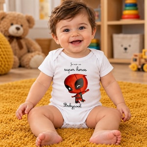 Könnte beinhalten: Ein weißer kurzärmeliger Babybody mit einer Grafik einer roten Superheldenfigur. Der Text über der Figur lautet "Je suis un super heros" und der Text unter der Figur lautet "Babypool" in einer schwarzen Schreibschrift.