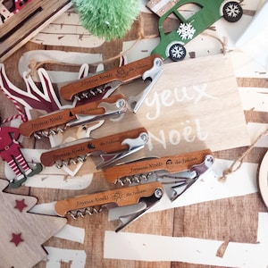 Op de afbeelding: Een verzameling houten kurkentrekkers met metalen accenten, elk gegraveerd met "Joyeux Noël" en "de Julien". De afbeelding bevat ook kerstversieringen zoals een houten boom, een rendierornament en een groene auto.