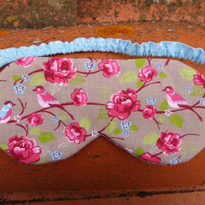 May include: A gray sleep mask with a floral pattern featuring pink roses and blue birds. The mask has a light blue elastic band.