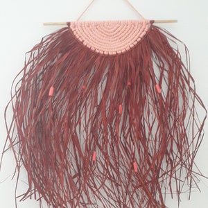 Peut inclure: Une suspension murale en macramé rouge et rose avec une frange de longues mèches fines. La suspension est décorée de petites perles roses.
