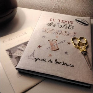 Pode incluir: Um diário branco coberto de linho com o título "LE TEMPS des FILS" e o texto "agenda de brodeuse" bordado em fio castanho. A capa também apresenta uma pequena bobina de fio, uma agulha e um par de tesouras douradas.