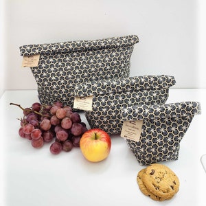 Könnte beinhalten: Drei Stoffaufbewahrungstaschen mit einem schwarz-goldenen geometrischen Muster. Ein Bund roter Trauben, ein rot-gelber Apfel und zwei Schokoladen-Chip-Cookies liegen neben den Taschen. Jede Tasche hat ein kleines Etikett.