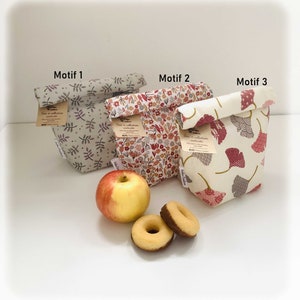Peut inclure: Trois sacs à déjeuner réutilisables avec différents motifs floraux. Les sacs sont pliés et portent une étiquette indiquant "Sac à collation lavable". Les sacs sont sur une surface blanche avec une pomme et deux beignets.