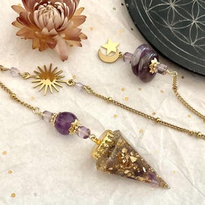 Peut inclure: Un pendule composé d'une pointe conique en résine remplie de flocons dorés et d'améthyste violette, fixé à une chaîne dorée. La chaîne comprend un pendentif soleil, un pendentif lune et étoile, ainsi que des perles violettes.