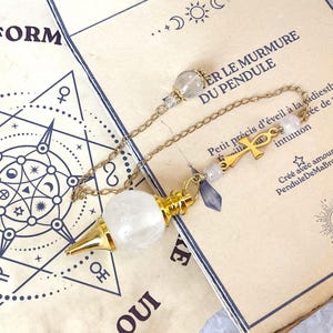 Könnte beinhalten: Ein Pendel mit einer klaren, kugelförmigen Kristallperle, die an einer goldfarbenen Metallspitze befestigt ist. Die Kette enthält kleine klare Perlen, ein goldfarbenes Ankh-Symbol und einen kleinen violetten Kristall, platziert auf einer Papierkarte.