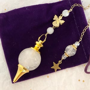 May include: A pendulum featuring a spherical white stone bead with a gold-toned metal cap and pointed tip. The gold-toned chain includes a small metal angel charm, two translucent round beads, and a dangling gold-toned star charm at the end.