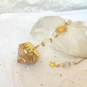 Peut inclure: Un pendule composé d'une pointe en résine facettée à éclats dorés, surmontée d'une coupelle en métal doré. La chaîne comporte des perles transparentes, un pendentif soleil et une étoile dorée. L'objet repose sur un cristal de quartz brut.
