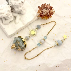 Könnte beinhalten: Ein Pendel mit einer facettierten, hellblauen Harzspitze mit Blattgoldflocken und einer goldfarbenen Kappe. Die Kette enthält einen goldenen Sonnenanhänger, hellblaue Perlen und einen kleinen goldenen Sternanhänger am Ende der Kette.