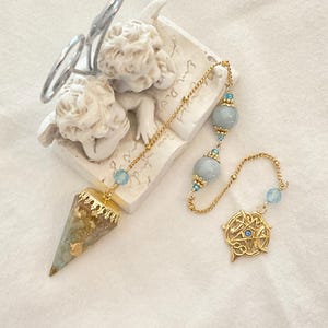 May include: A pendulum featuring a cone-shaped stone with gold-colored flakes, attached to a gold-colored chain with light blue beads and a circular gold-colored charm containing a pentagram and a small blue stone, resting near a cherub figurine.