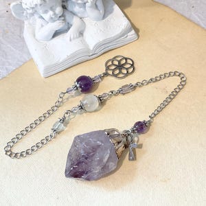 May include: A silver-toned metal chain pendulum featuring a large, raw, faceted purple amethyst point at the base. The chain is adorned with smaller clear and purple spherical beads, a small silver ankh charm, and a circular flower-like metal charm.