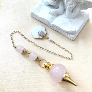 Peut inclure: Un pendule composé d'une grosse pierre de quartz rose sphérique fixée dans un capuchon et une pointe en métal doré. La chaîne dorée comporte deux perles de quartz rose plus petites et une breloque en forme de coquillage en émail blanc.