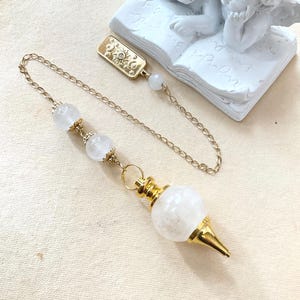 May include: A gold-toned metal pendulum featuring a white spherical stone, two smaller white beads, and a gold chain. The chain ends in a rectangular gold charm embossed with celestial sun and moon motifs, accented by a small white bead.