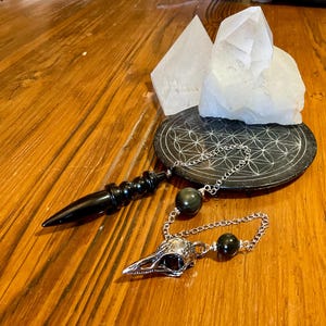 Könnte beinhalten: Ein schwarzes Obsidian-Pendel mit spitzer Form, befestigt an einer silberfarbenen Metallkette. Die Kette ist mit zwei runden Obsidianperlen und einem silberfarbenen Metallanhänger in Form eines Vogelschädels versehen. Das Pendel liegt neben einer schwarzen runden Scheibe mit weißem Lebensblumenmuster und zwei klaren Quarzkristallspitzen.