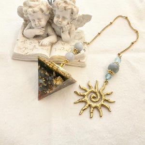 May include: A gold-colored sun-shaped pendant with a spiral center and radiating points, connected by a gold chain to a triangle-shaped pendant with gold flecks and blue beads. The sun pendant is approximately 2 inches in diameter.