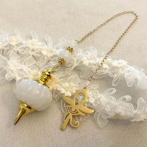 Op de afbeelding: Een wit en goudkleurig pendel met een gouden ketting en een Ankh-bedel. De pendel heeft een witte, afgeronde behuizing en een gouden punt. De ketting is bevestigd aan een witte kant en bloemenrand.