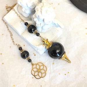 May include: A pendulum featuring a large, smooth, black spherical stone with a gold-toned metal cap and pointed tip. The chain includes smaller black beads with gold-toned accents and a circular gold-toned metal charm with a floral pattern.