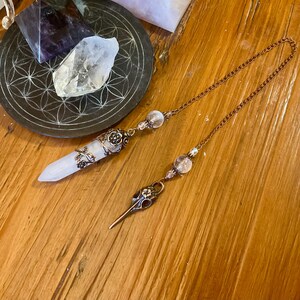 Könnte beinhalten: Ein kupferfarbenes Kettenpendel mit einem spitzen weißen Kristall in einer floralen Metallkappe, verbunden mit einem Anhänger in Form eines Vogelschädels, verziert mit kleinen klaren Perlen entlang der metallischen Kette.