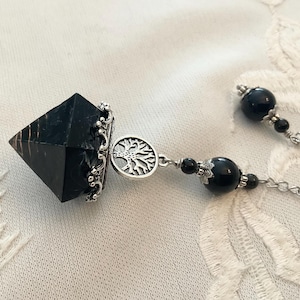 May include: A black crystal pendulum with a silver-coloured tree of life charm and chain. The pendulum has a faceted, pyramid shape. The chain features black beads and a decorative silver-coloured cap.