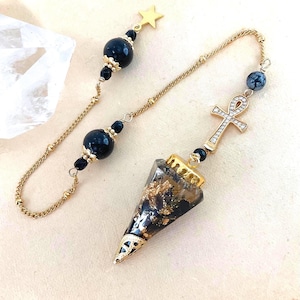 May include: A pendulum featuring a cone-shaped black and gold resin point with a gold metal cap, attached to a gold chain. The chain includes two black beads, a small gold star charm, and a gold ankh symbol encrusted with clear crystals.