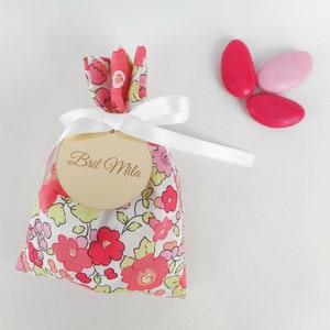 May include: A small floral fabric bag tied with a white ribbon and a wooden tag that reads "Brit Mila". Three pink sweets are next to the bag. The bag has a pink, red, and yellow floral pattern.