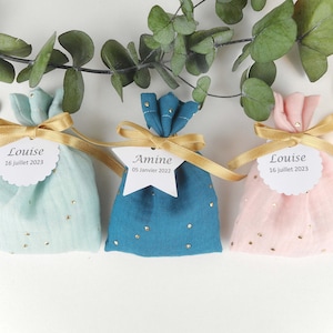 Puede incluir: Tres pequeñas bolsas de regalo de tela con cinta dorada y etiquetas personalizadas. Las bolsas son azul claro, azul verdoso y rosa con puntos de purpurina dorada. Las etiquetas tienen los nombres "Louise", "Amina" y "Louise" con fechas.