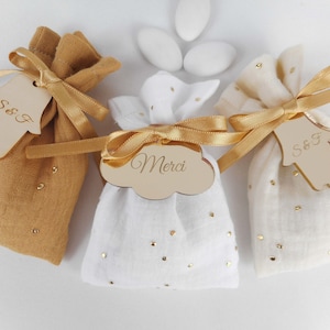 以下が含まれることがあります： ベージュ、白、オフホワイトの3色の小さな布製ギフトバッグ。金色のリボンとタグで飾られています。バッグには金のドットが装飾されています。1つのタグには「Merci」と書かれています。