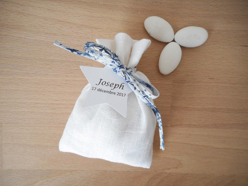 May include: White linen favor bag with a blue and white patterned ribbon tied around it. A white star-shaped tag is attached to the ribbon with the text "Joseph 17 d&eacute;cembre 2017".