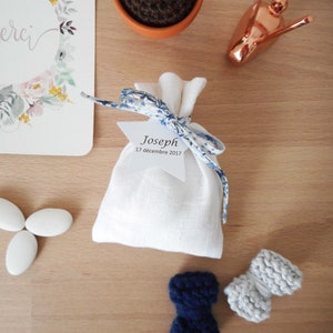 May include: A white linen bag with a blue and white ribbon tied around it. The bag has a white star-shaped tag with the text "Joseph 17 d&eacute;cembre 2017" on it. There are also two small knitted bows, one blue and one gray, on a wooden surface.