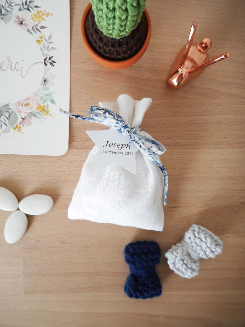 May include: A white linen bag with a blue and white ribbon tied around it. The bag has a white star-shaped tag with the text "Joseph 17 d&eacute;cembre 2017" on it. There are also two small knitted bows, one blue and one gray, on a wooden surface.