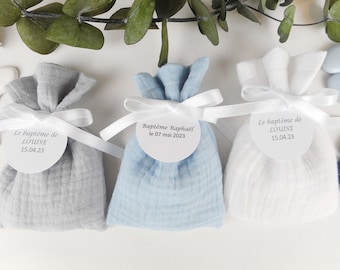 May include: Three small fabric gift bags with white satin ribbon ties. The bags are grey, light blue, and white. Each bag has a round white label with black text. The labels read "Le baptême de LOUISE 15.04.23", "Baptême Raphaël le 07 mai 2023", and "Le baptême de LOUISE 15.04.23".
