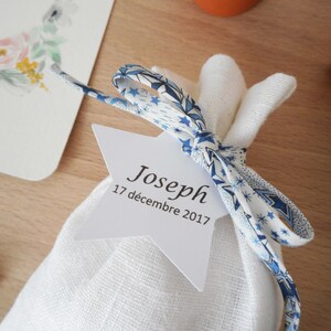 May include: A white fabric bag with a blue and white patterned ribbon tied around it. A white star-shaped tag is attached to the ribbon with the text "Joseph 17 d&eacute;cembre 2017".