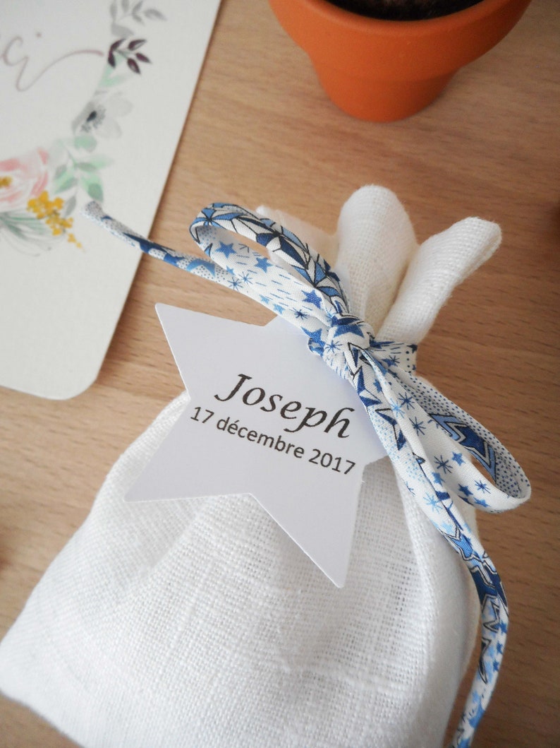 May include: A white fabric bag with a blue and white patterned ribbon tied around it. A white star-shaped tag is attached to the ribbon with the text "Joseph 17 d&eacute;cembre 2017".