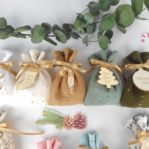 May include: A set of six small fabric gift bags with gold ribbon and wooden tags. The tags feature various Christmas-themed designs, including a gingerbread man, a stocking, a candy cane, a Christmas tree, and a star. The bags are filled with small gifts and are perfect for a baby's first Christmas.