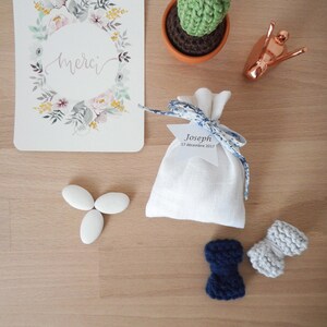 May include: A white favor bag with blue and white ribbon and a tag that says "Joseph 17 decembre 2017". There are three white almond candies, a crocheted cactus in a terracotta pot, a copper snail figurine, and two crocheted bow ties, one blue and one gray.