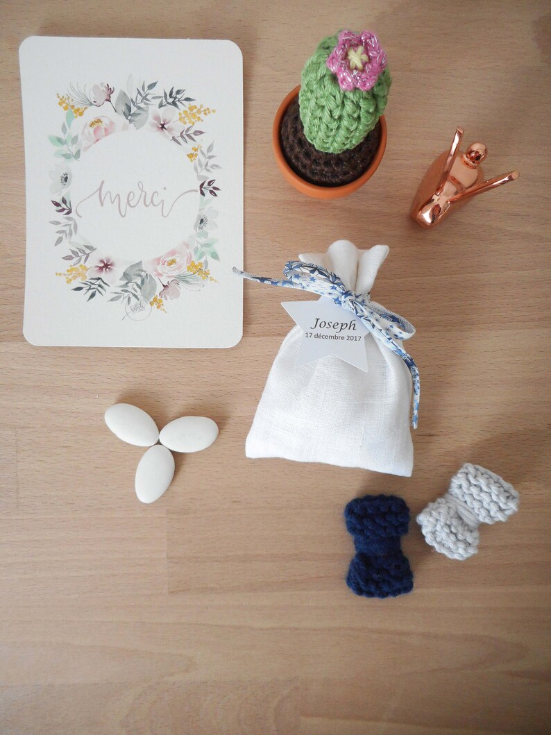 May include: A white favor bag with blue and white ribbon and a tag that says "Joseph 17 decembre 2017". There are three white almond candies, a crocheted cactus in a terracotta pot, a copper snail figurine, and two crocheted bow ties, one blue and one gray.