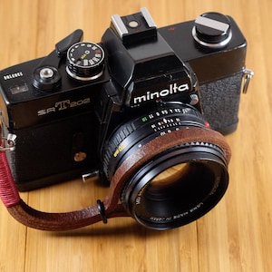 Puede incluir: Una cámara de película negra Minolta SR-T 202 con una correa de cuero marrón. La cámara tiene un objetivo negro con un anillo plateado y marcas blancas.