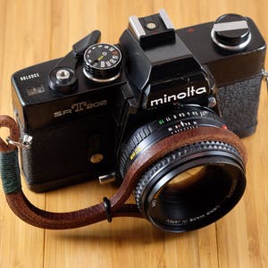 May include: Black Minolta SR-T 202 film camera with a brown leather strap and lens. The camera has a silver flash attachment and a black lens with brown leather trim. The camera is on a light brown wooden surface.