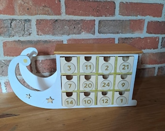 Puede incluir: Trineo de madera blanco con la parte superior pintada de oro. El trineo tiene 12 cajones numerados, cada uno con un círculo dorado que contiene un número del 1 al 24. Los cajones están dispuestos en cuatro filas de tres.