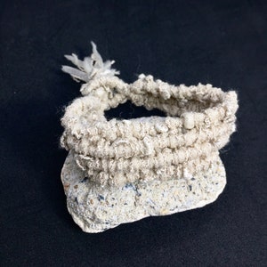 Puede incluir: Un brazalete blanco de ganchillo con un borla, descansando sobre una roca gris clara.