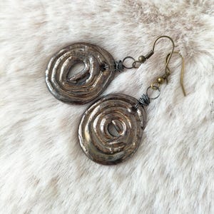 Può includere: Un paio di orecchini argentati metallici fatti a mano. Ogni orecchino presenta un design a spirale con una superficie strutturata e una chiusura a gancio. Gli orecchini sono esposti su una superficie bianca e soffice.