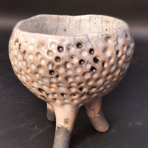 Peut inclure: Petit bol en céramique fait à la main, avec une surface texturée et trois petits pieds. Le bol est blanc cassé avec des taches sombres et un glaçage craquelé, lui donnant un aspect rustique. Les pieds sont gris-brun foncé à la base.