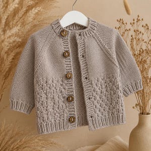 Könnte beinhalten: Eine hellbeige, langärmelige Strickjacke mit einem strukturierten Korbgeflechtmuster auf den unteren Vorderteilen, einem gerippten Rundhalsausschnitt, gerippten Bündchen und einer vertikalen Reihe aus fünf runden braunen Holzknöpfen.