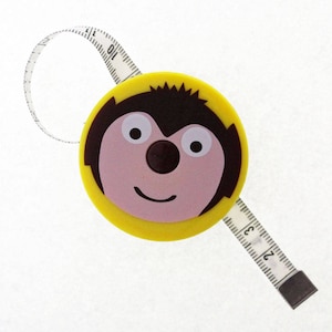 Può includere: Nastro di misura retrattile giallo con un viso di animale dei cartoni animati. Il viso ha i capelli castani, gli occhi neri, un naso rosa e un sorriso. Il nastro di misura ha i numeri 10, 11, 12, 21, 22 e 23 stampati.