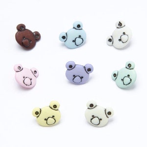 Può includere: Assortimento di otto bottoni di plastica a forma di orso di diversi colori. I bottoni sono di vari colori pastello, tra cui blu, rosa, viola, verde, giallo e marrone. Ogni bottone presenta un semplice contorno nero di un viso di orso.